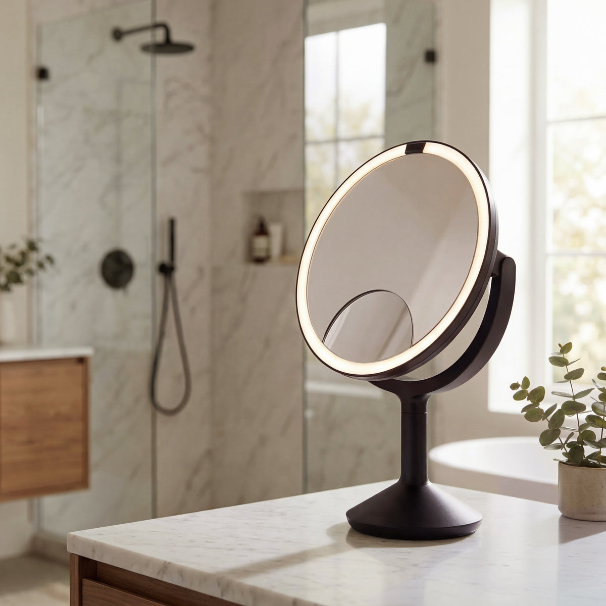Simplehuman's Sensor Mirror Trio Max in Matte Black in a bathroom setting with shower and plants in the background.